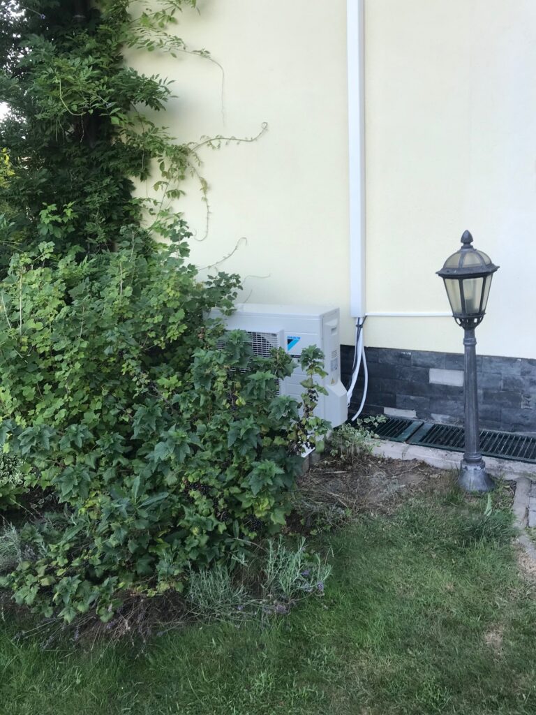 Venkovní jednotka klimatizace Daikin u stěny domu obklopená zelení