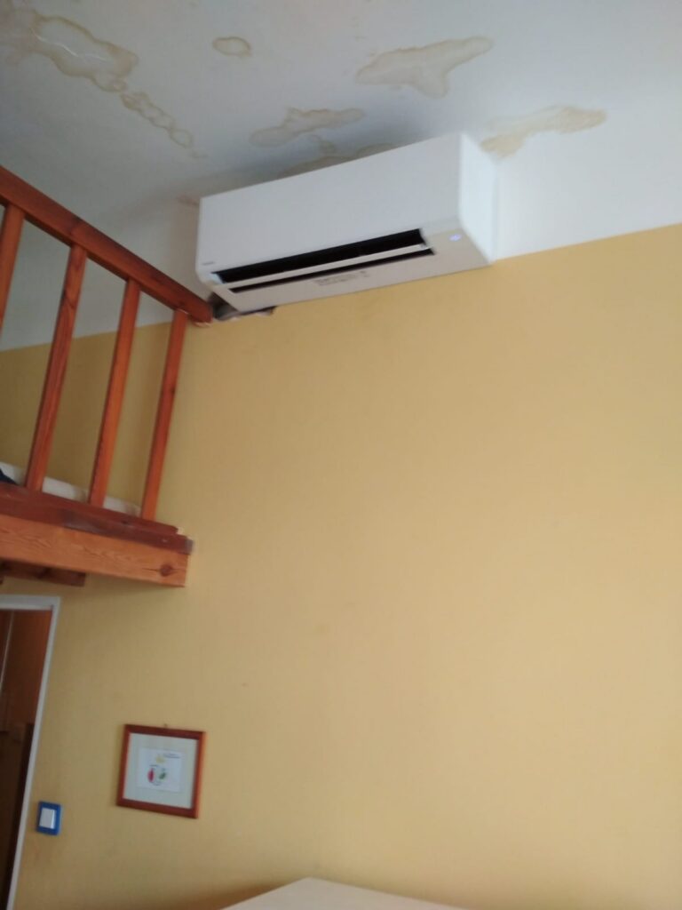 Bílá nástěnná klimatizace umístěná pod stropem vedle dřevěného schodiště